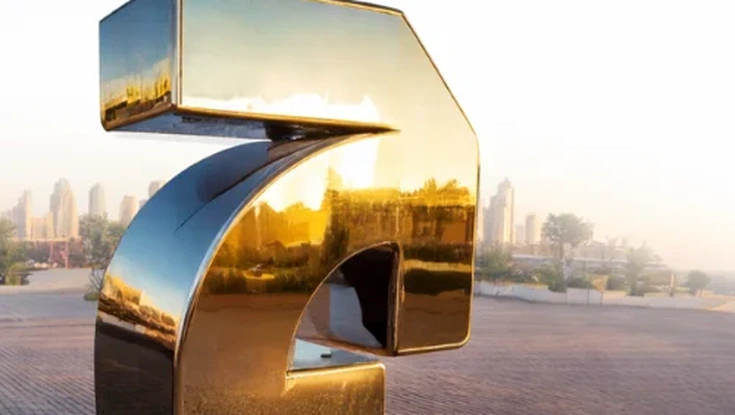 Reflective golden question mark sculpture in an outdoor urban setting at sunset.